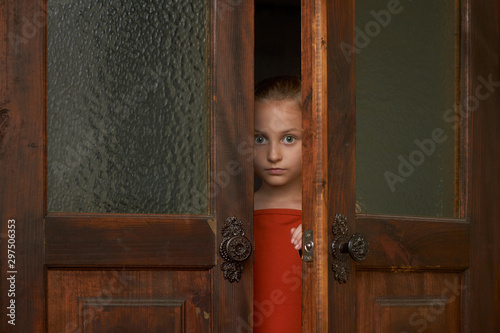 A little scared girl is looking through the door slit. A girl is watching someone or something through ajar door. photo