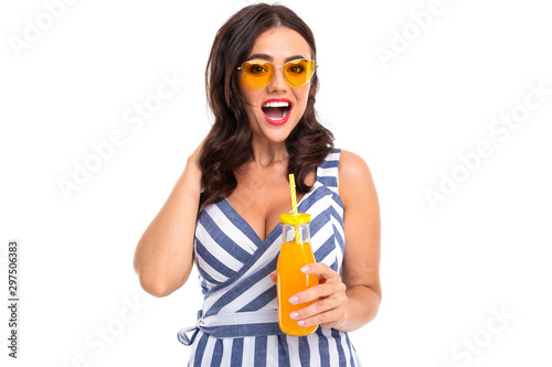 A young girl with a delightful smile, flat teeth, red lipstick, long wavy chestnut hair, beautiful makeup, in a white and blue dress in a stripe with a cleavage, yellow glasses holds orange juice in a