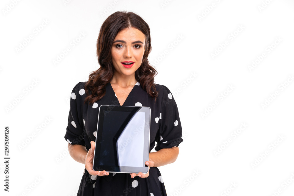 Young girl with delightful smile, flat teeth, red lipstick, long wavy chestnut hair, beautiful makeup, in black and white dress in peas holds a tablet Young girl with delightful smile, flat teeth, red lipstick, long wavy chestnut hair, beautiful makeup, in black and white dress in peas holds a tablet