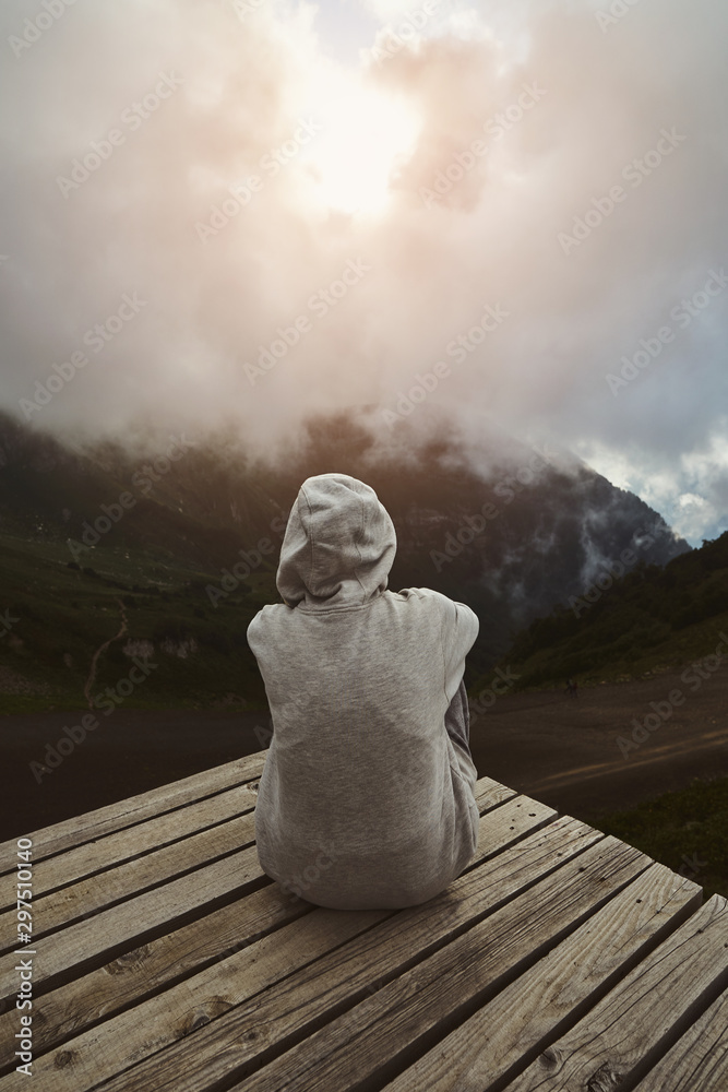 a girl in a grey hoodie sitting on a viewpoint and watching sunset in the mountains