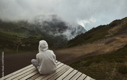 a girl in a grey hoodie sitting on a viewpoint in the mountains