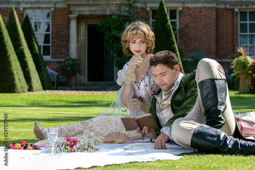 Young lovers dressed in vintage clothing sitting on picnic blanket. Gentleman reading to his lover from a book of poems