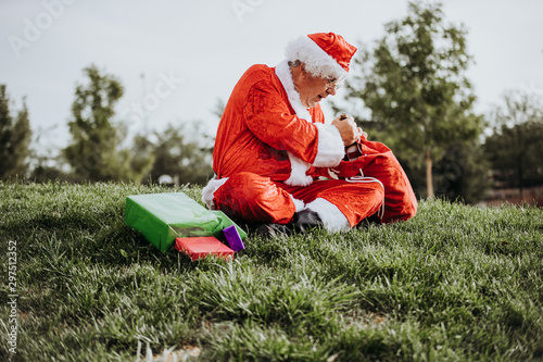 Vertical stock photo of Santa Claus without beard crouched on the floor organizing gifts with his red bag. Christmas time