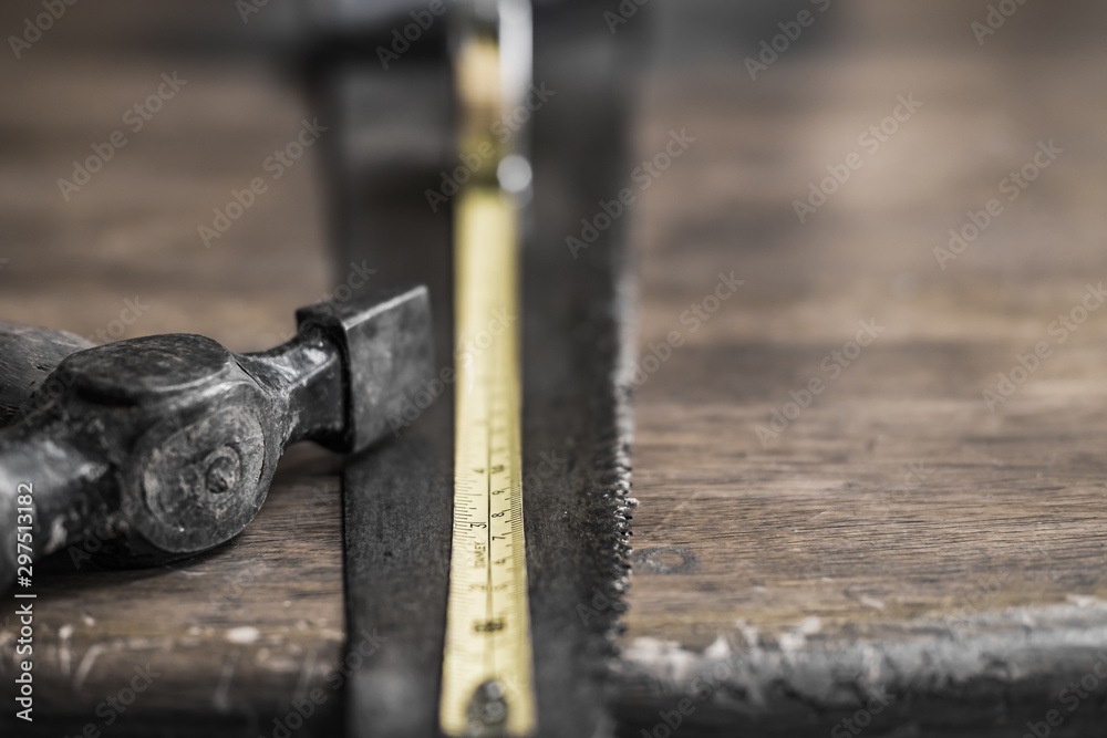 Close-up, shallow focus of an extended measuring tape showing the scales. Also are an old hammer and wood saw seen on an old, wooden workshop table. Close-up, shallow focus of an extended measuring tape showing the scales. Also are an old hammer and wood saw seen on an old, wooden workshop table.