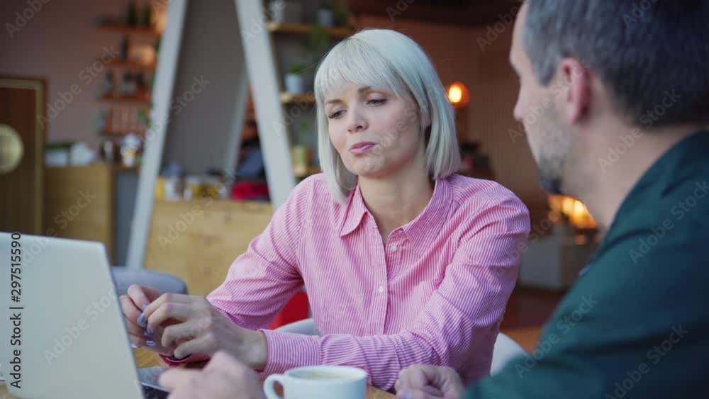 Camera panning left to confident female entrepreneur in pink shirt discussing data from laptop with male colleague while sitting at table in cafe