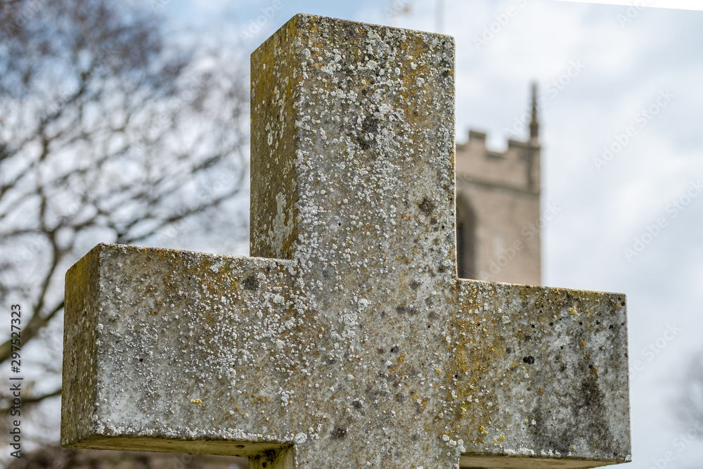 Shallow focus, showing the stone texture of a crucifix shaped gravestone. The background shows a typical, English church tower.
