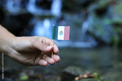 National flag of Mexico on wooden stick. Young boy holds national athem near waterfall on stream in autumn times. Concept of humanity and nature. United Mexican States in hand. Flag for packet