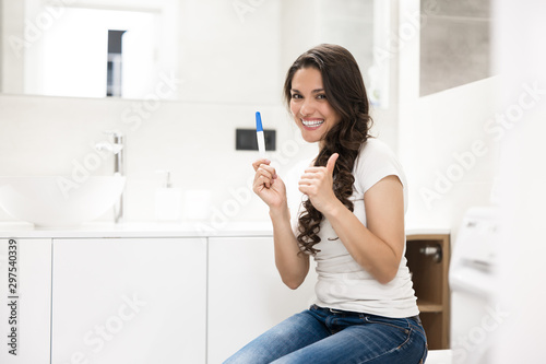 young beautiful woman sits on the toilet bowl holding pregnancy test looking happy showing like sign
