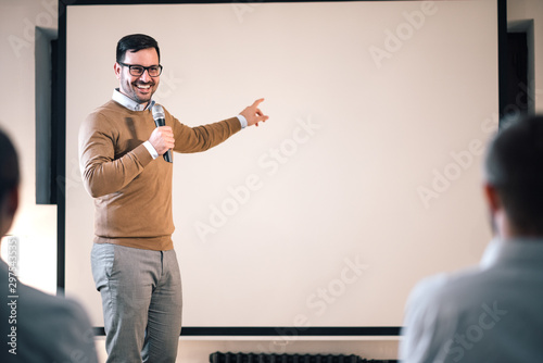 Stampa su tela Portrait of a speaker on a seminar, talking on microphone and pointing at blank screen