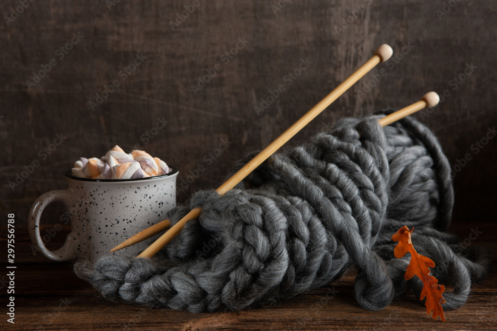 Knitting with thick gray threads on wooden knitting needles and a mug with a hot drink and marshmallows. Still life in vintage style