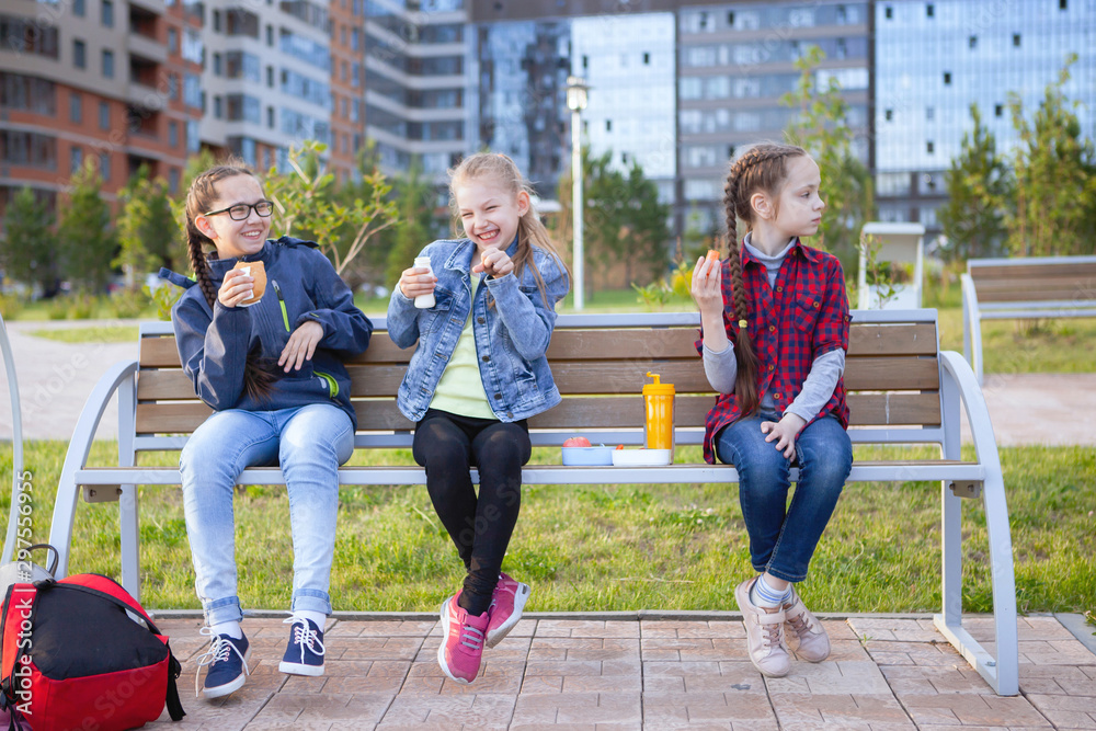 Teen girls eat on a bench in a city park.