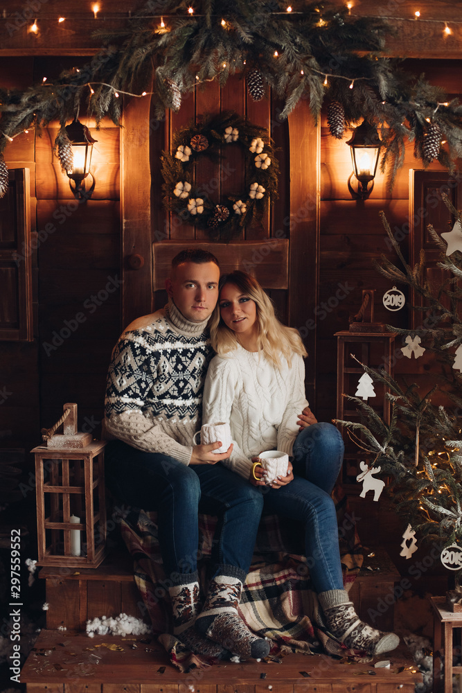 Christmas portrait of a romantic couple. They sit by a beautiful house and have hot coffee. New Year mood of lovers on the background of a wooden house, with fans. Close - up