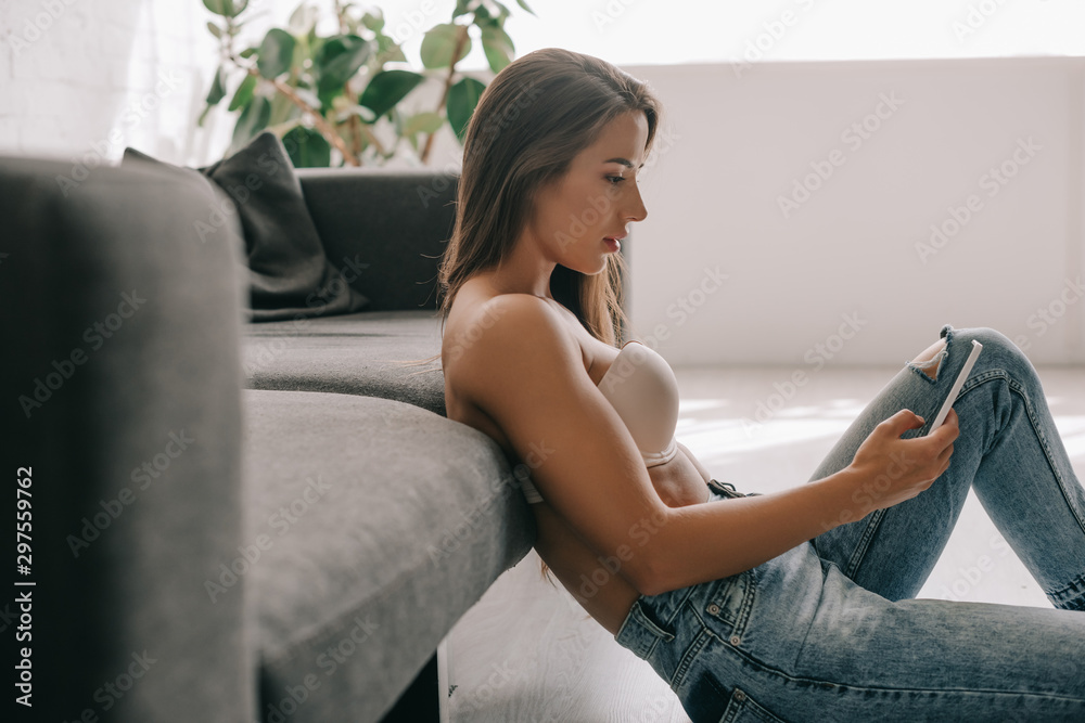 beautiful girl in white bra and jeans using smartphone while sitting near sofa beautiful girl in white bra and jeans using smartphone while sitting near sofa