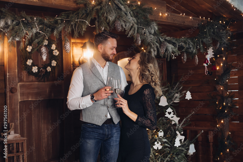 Portrait of happy young couple in elegant outfits smiling face to face with two glasses of champagne. They standing in beautifully decorated room at Christmas. They are hugging and smiling.