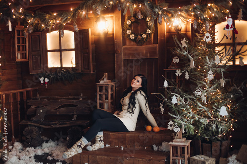 Portrait of cheerful and positive woman with long dark hair in sweater, jeans and warm socks holding wrapped gift for Christmas while sitting under decorated Christmas tree and snowfall. © Вячеслав Косько