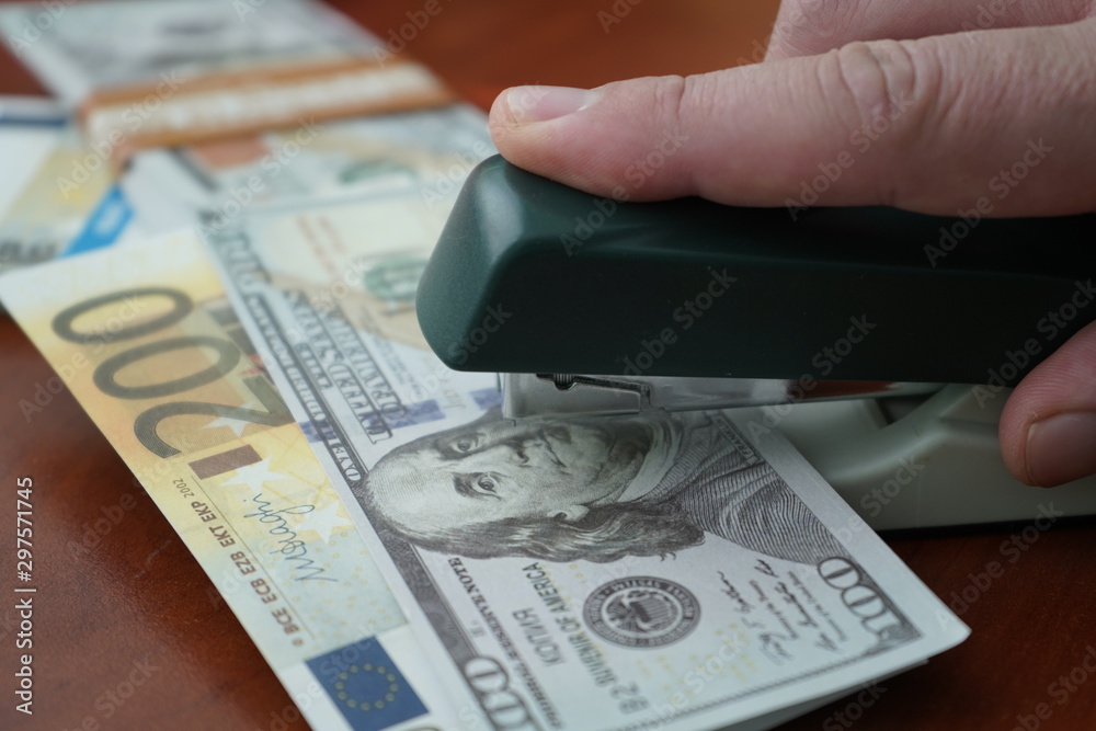 The stapler fastens the dollar and euro notes with iron clips. Stapler with paper clips on the background of banknotes. The stapler fastens the dollar and euro notes with iron clips. Stapler with paper clips on the background of banknotes.