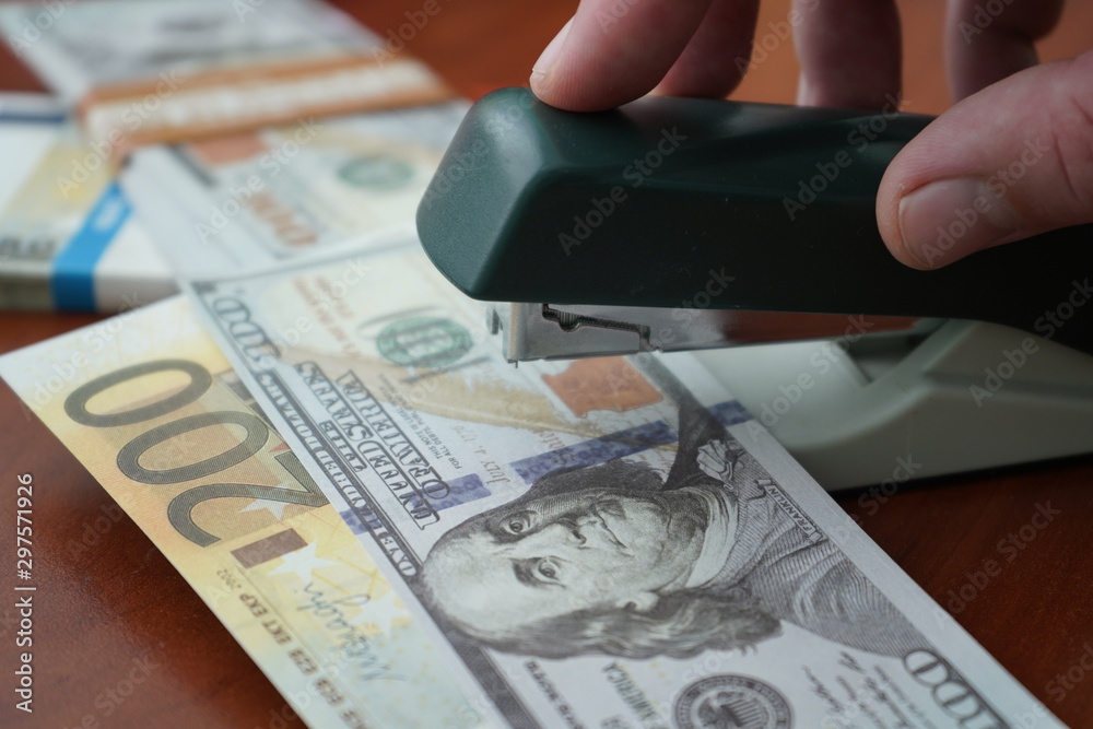 The stapler fastens the dollar and euro notes with iron clips. Stapler with paper clips on the background of banknotes. The stapler fastens the dollar and euro notes with iron clips. Stapler with paper clips on the background of banknotes.
