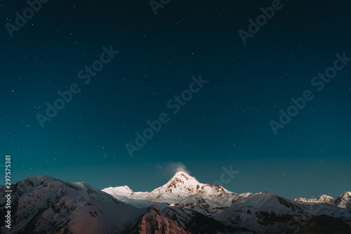 Georgia. Winter Night Starry Sky With Glowing Stars Over Peak Of Mount Kazbek Covered With Snow. Beautiful Night Georgian Winter Landscape