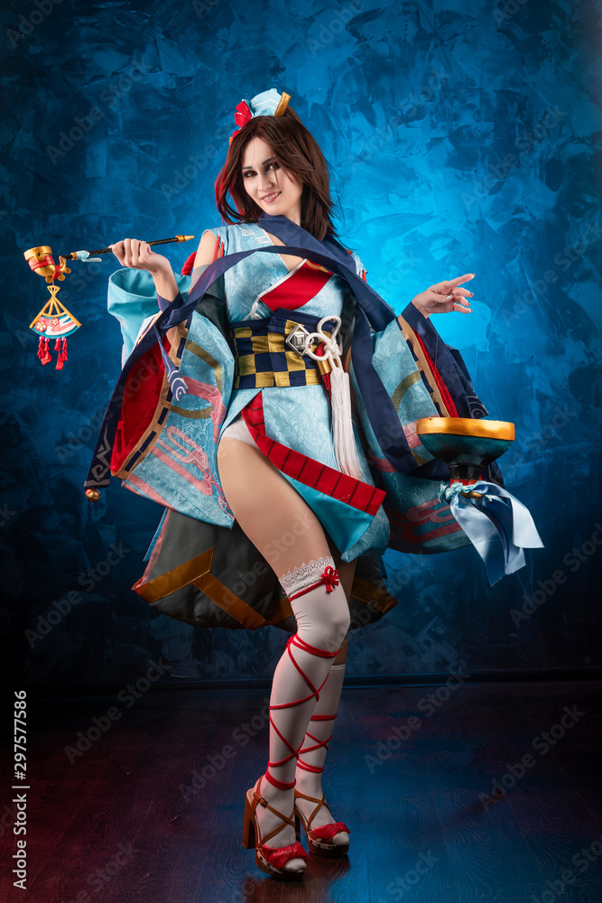Beautiful leggy busty cosplayer girl wearing a stylized Japanese kimono  costume cheerfully posing holding a fake pipe on a blue background.  Stock-Foto | Adobe Stock