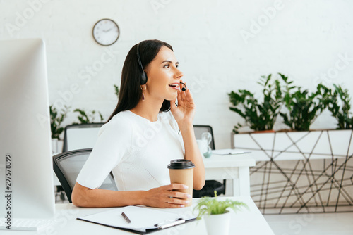 happy broker touching headset while holding coffee to go in office