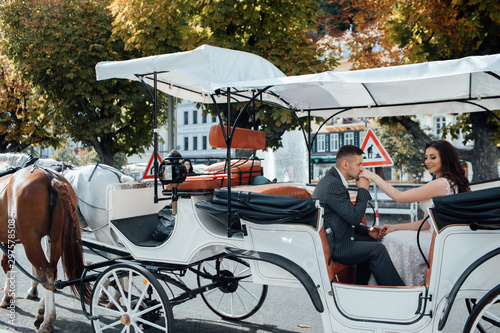 Happy couple sits are in coach in city. Guy kisses girl's hand. Beautiful couple enjoying time spending with each other. Fashion photo. Handsome guy in suit and girl in white dress. Couple in love.