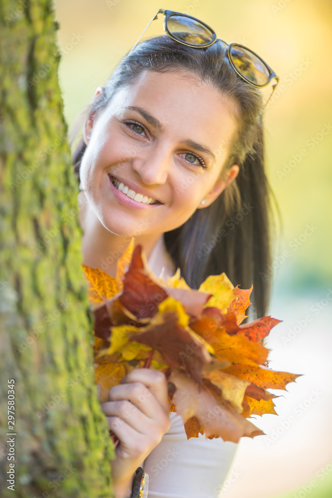 Happiness woman sitting in autumn park holding bouquet with fall leaves cute look from behind a tree Happiness woman sitting in autumn park holding bouquet with fall leaves cute look from behind a tree