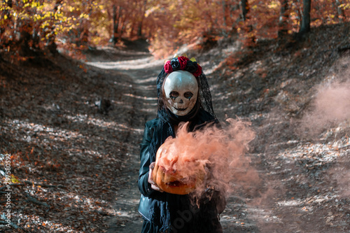 Fototapeta Naklejka Na Ścianę i Meble -  Photo of  walking girl with creepy skull mask in the autumn woods, holding a carved smoky Halloween pumpkin.  On background path in autumn forest with fall leaves. Concept about Halloween.