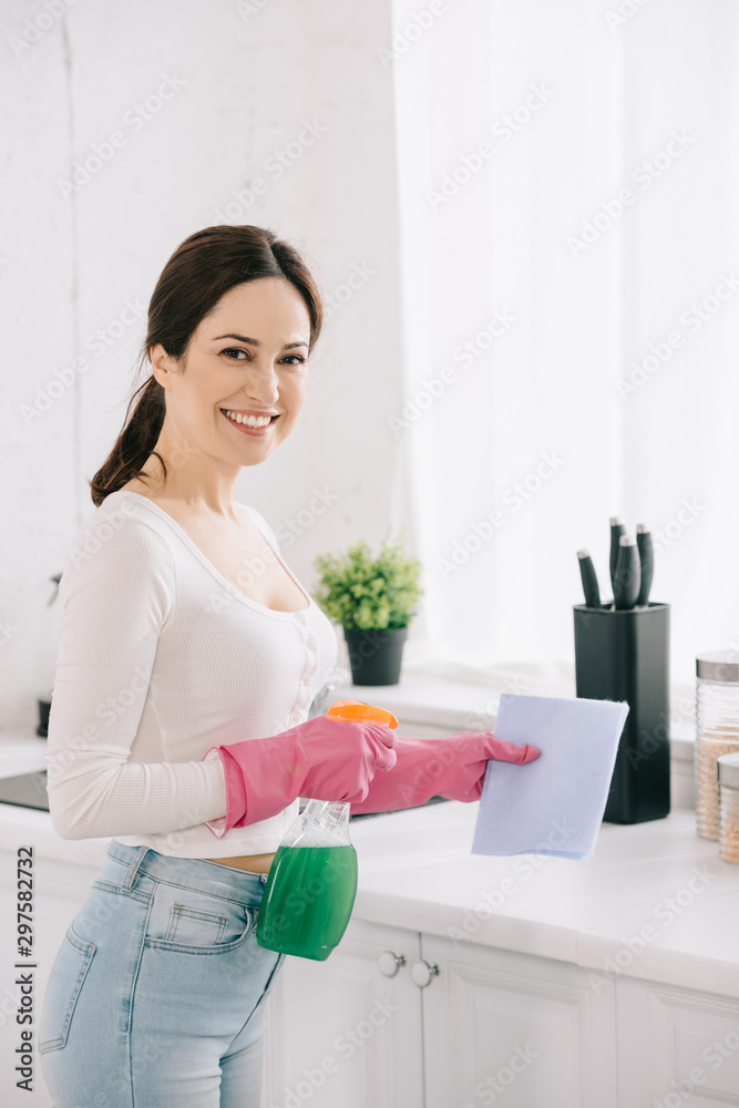 cheerful housewife looking at camera while holding spray bottle and rag in kitchen cheerful housewife looking at camera while holding spray bottle and rag in kitchen