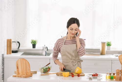 smiling woman in apron cutting bell pepper while standing near kitchen table with vegetables and talking on smartphone