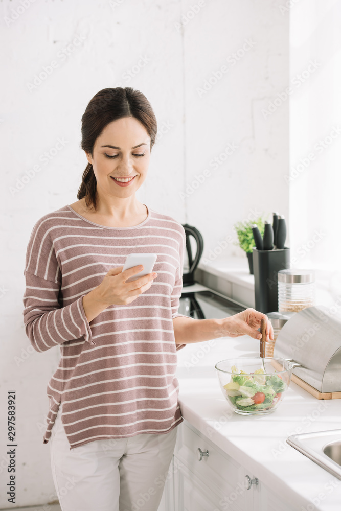 young, smiling woman using smartphone while standing at kitchen table near bowl with vegetable salad young, smiling woman using smartphone while standing at kitchen table near bowl with vegetable salad