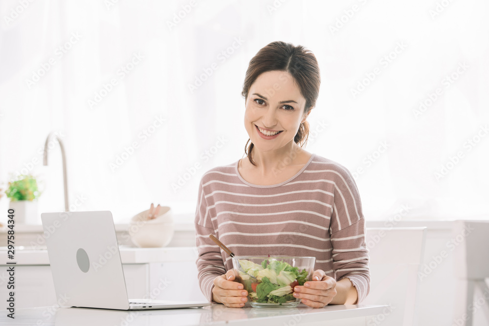 young, smiling woman looking at camera while sitting at table with bowl of vegetable salad near laptop