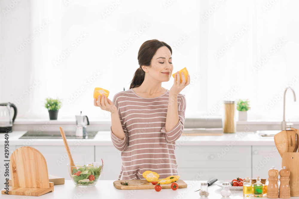 pretty, young woman smelling cut bell pepper while standing at kitchen table near fresh vegetables pretty, young woman smelling cut bell pepper while standing at kitchen table near fresh vegetables