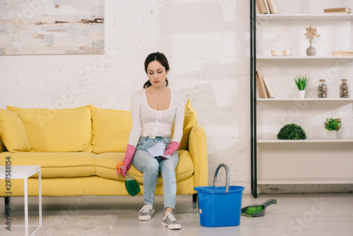 tired housewife sitting on yellow sofa and holding spray bottle near bucket, scoop and brush on floor