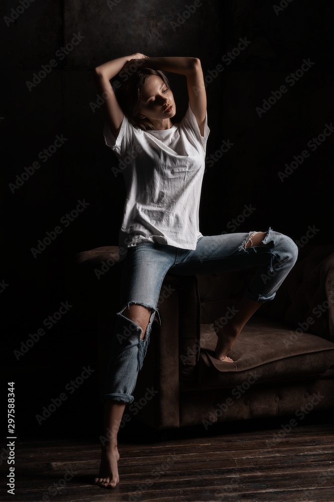 Attractive sexy fashion woman with brunette hairstyle in shirt and jeans sitting on armchair in loft room. Portrait of seductive hot stylish woman model on black background in studio