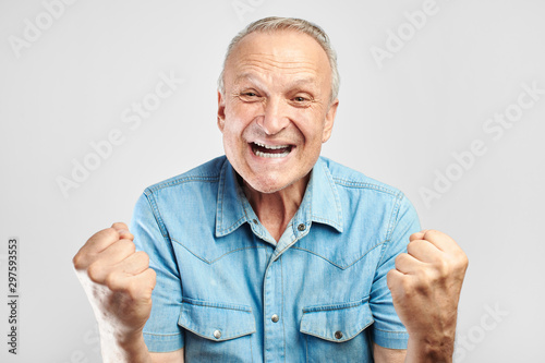 Positive gray-haired old man joyfully clenches fists, celebrates victory and success, smiles and enjoys the good news or sale isolated on white background photo