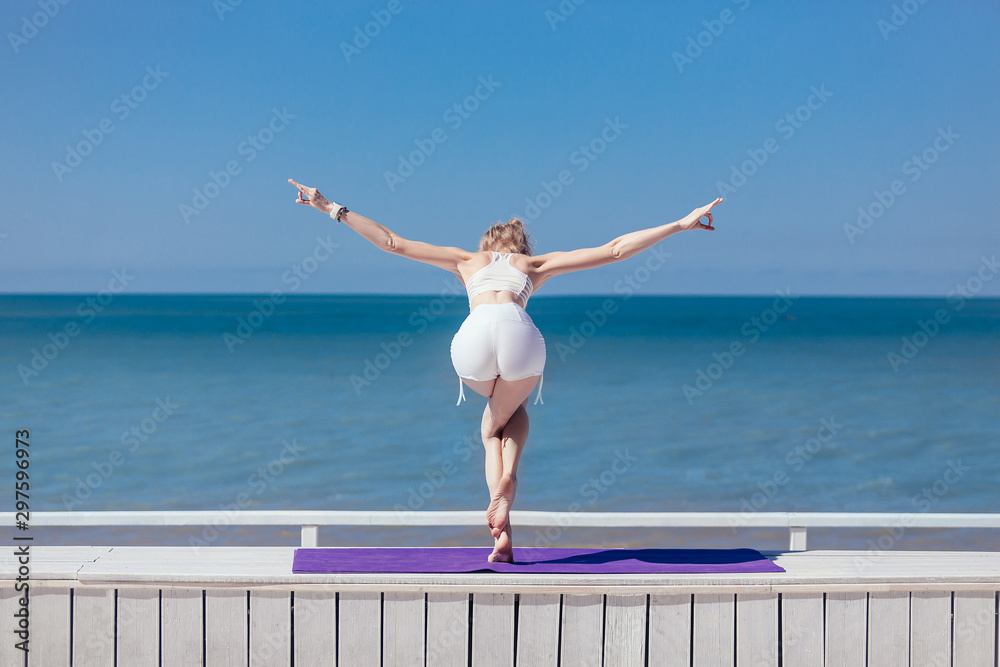 Obraz premium Sporty young woman doing stretching, standing Eagle Pose, Garudasana. Slim girl practicing yoga outdoor by sea or ocean, blue sky, white wooden terrace. Calm, relax, healthy lifestyle concept
