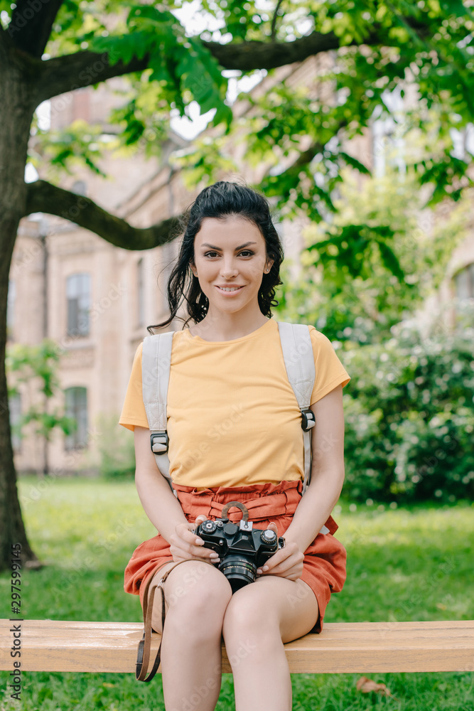 Happy girl holding digital camera while sitting on bench near university Happy girl holding digital camera while sitting on bench near university
