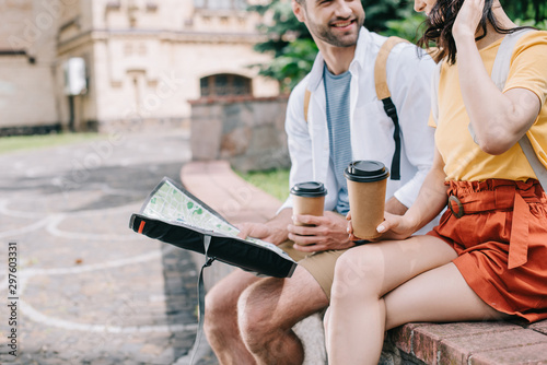 cropped view of happy bearded man with paper cup and map sitting near girl