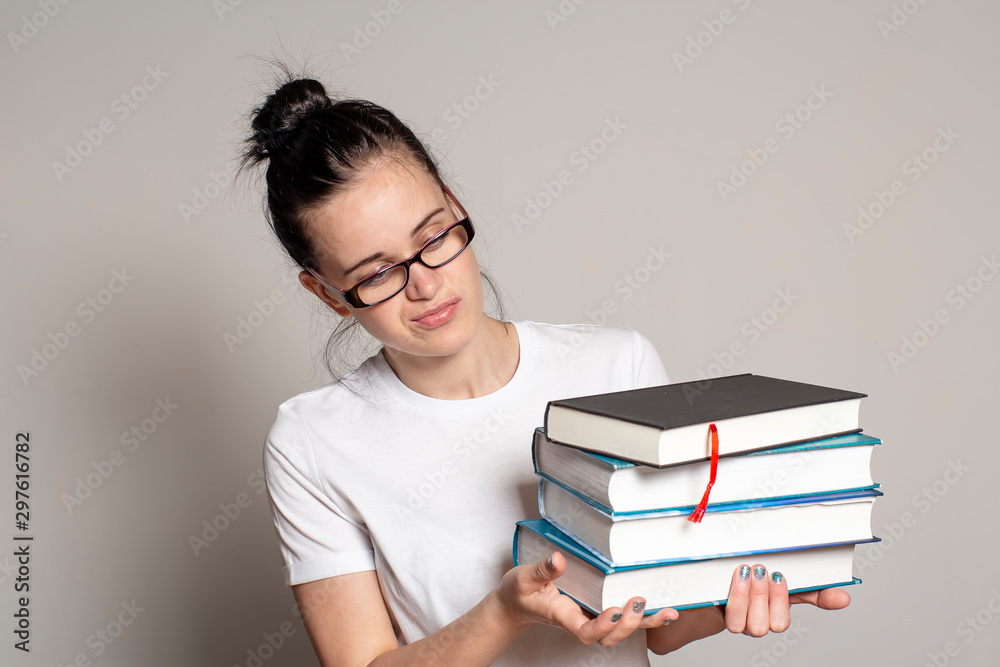 Pretty woman student with glasses holds a stack of heavy books in hands and looks at them puzzled. Pretty woman student with glasses holds a stack of heavy books in hands and looks at them puzzled.