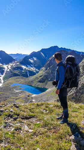 A young man with a big hiking backpack standing above a clear, navy blue lake hiding between tall mountain peaks. Some of the slopes are covered with snow. In the back is another mountain range.