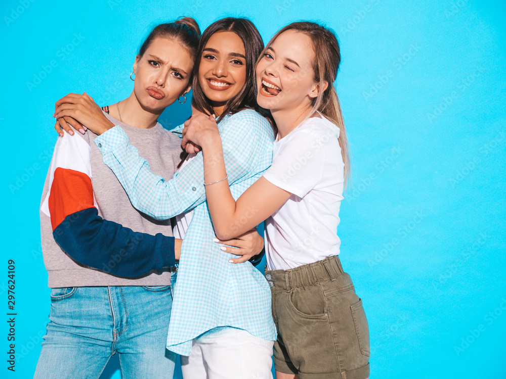 Three young beautiful smiling hipster girls in trendy summer clothes. Sexy carefree women posing near blue wall in studio. Positive models having fun and hugging Three young beautiful smiling hipster girls in trendy summer clothes. Sexy carefree women posing near blue wall in studio. Positive models having fun and hugging