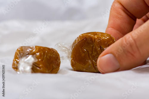 Close up of light brown caramel sweets in plastic wrap. Hand picking up one candy. Two caramel candies on white paper. Selective focus. Sweet treat.