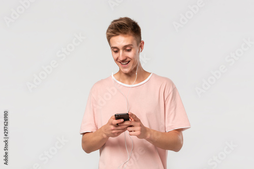 Smiling blond teenager in a pink tee listening music on a smartphone with wired headphones isolated over light grey background.