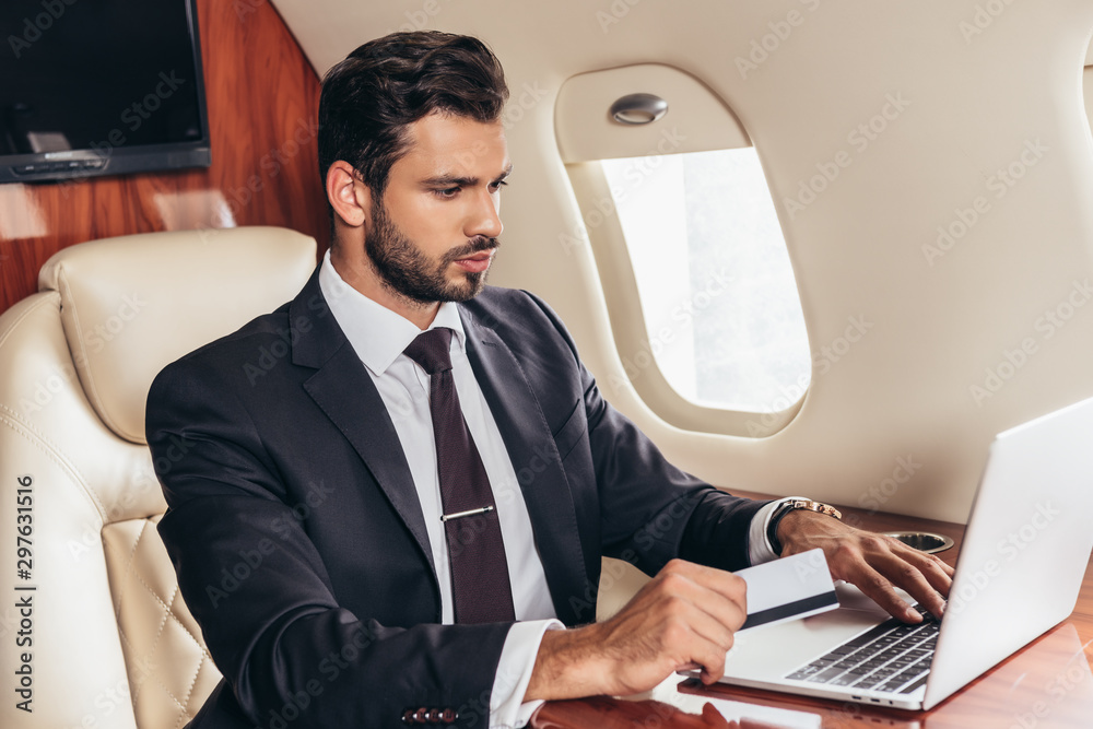 Fototapeta premium handsome businessman in suit holding credit card and using laptop in private plane