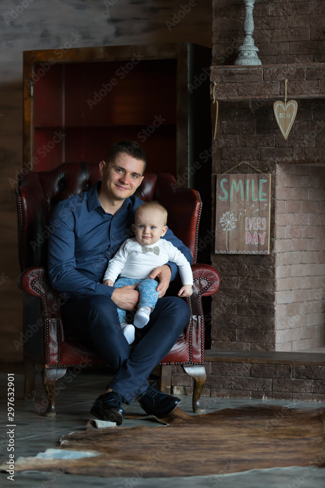 A father sits in a chair and holds his little son in his lap. They are both smiling. Family life. A father sits in a chair and holds his little son in his lap. They are both smiling. Family life.