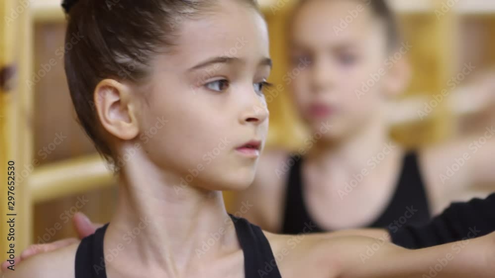 Close up faces of concentrated young girls of primary school age standing by wall bars and practicing ballet movements, handheld