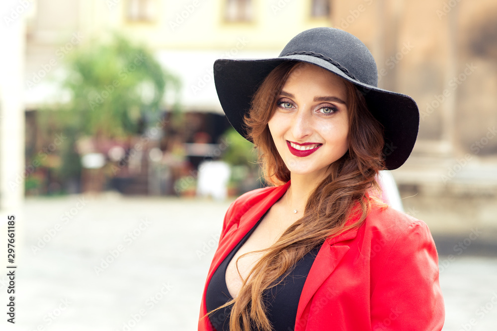 Fashionable young woman is wearing black hat looking at camera outdoors. Outdoor portrait of a young happy lady posing on a street. Copy space.