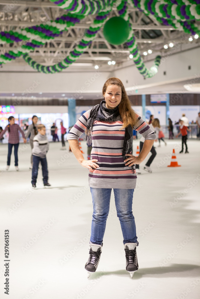 Obraz premium Portrait of cheerful woman dressed sweater, jeans and scarf standing on ice in hockey skates, indoor ice-scating rink with people