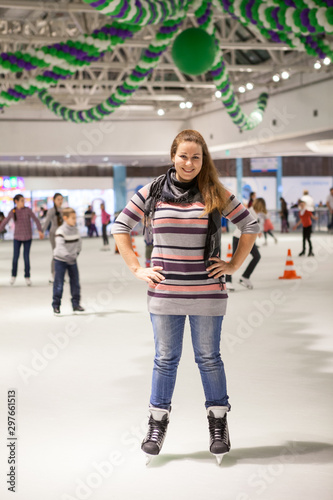Portrait of cheerful woman dressed sweater, jeans and scarf standing on ice in hockey skates, indoor ice-scating rink with people © Kekyalyaynen