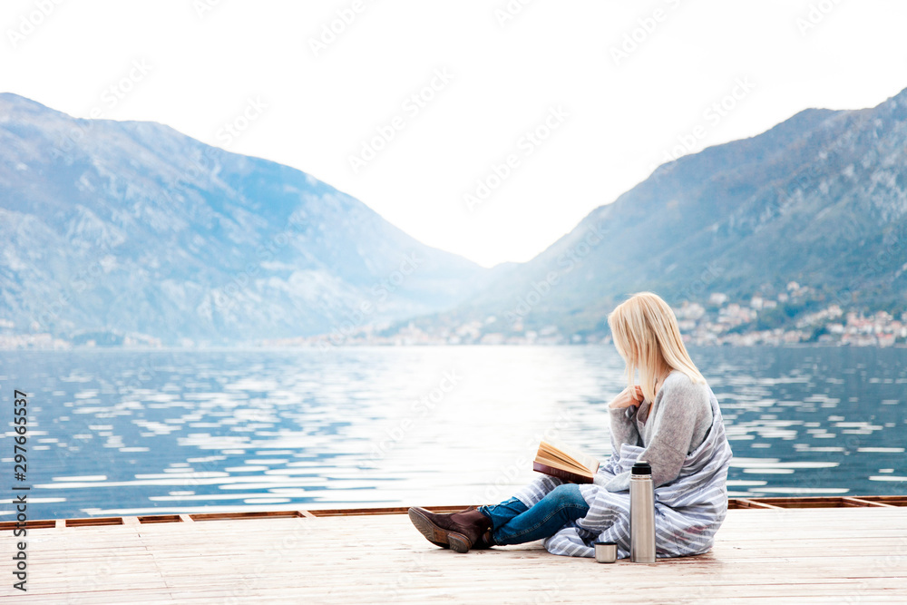 Woman is reading on wooden pier by winter sea, beach, mountains. Cozy picnic with coffee, hot beverages, tea in thermos and mug, warm plaid, opened book. Girl is enjoying nature, wellbeing.